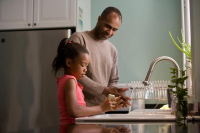 صورة أحد الوالدين يساعد طفلًا على غسل أيديهم في حوض المطبخ.
